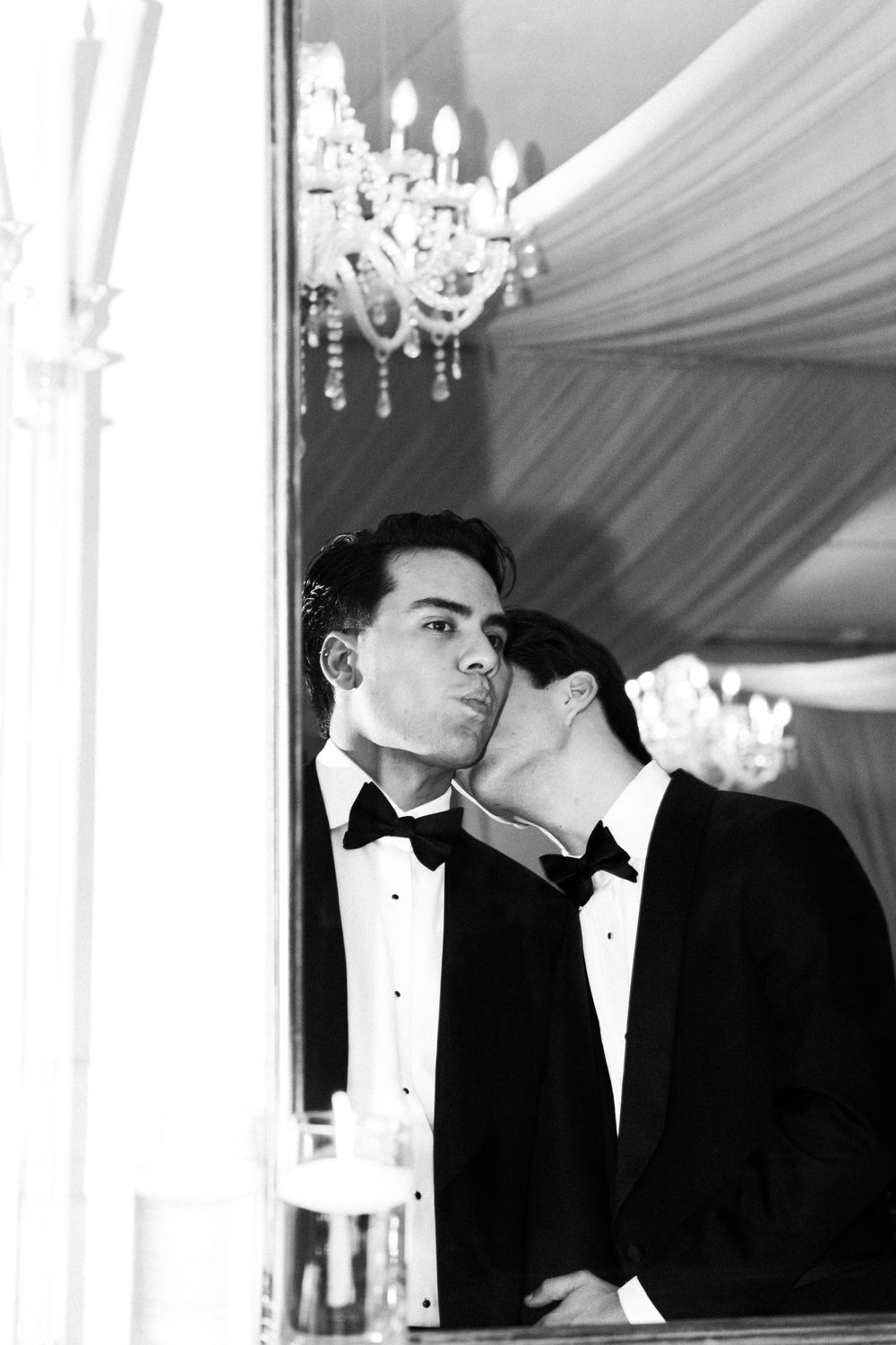 A black and white portrait of two people in formal attire with tuxedos under elegant chandeliers at an upscale event.