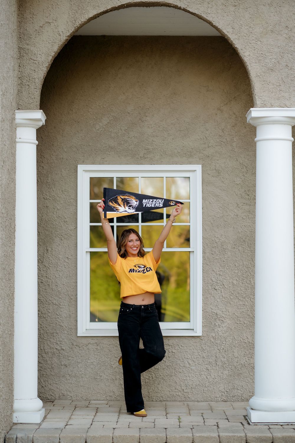 Smiling young woman holding a Mizzou Tigers pennant under an archway wearing a gold Missouri Tigers crop top.