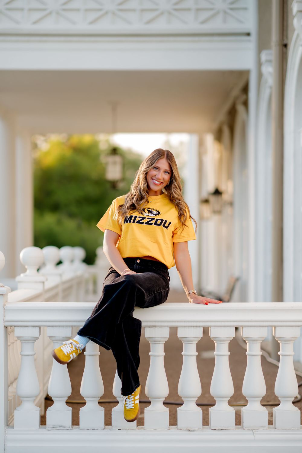 Young woman in Mizzou yellow t-shirt sits smiling on white balustrade railing of elegant porch.