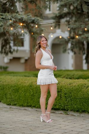 Woman in white mini dress and heels smiling outdoors near hedges with string lights and brick building.
