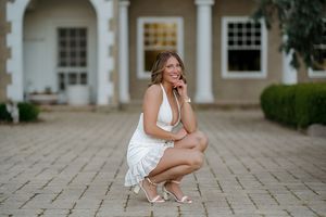 A smiling woman in a white dress crouches on a brick driveway in front of an elegant home.