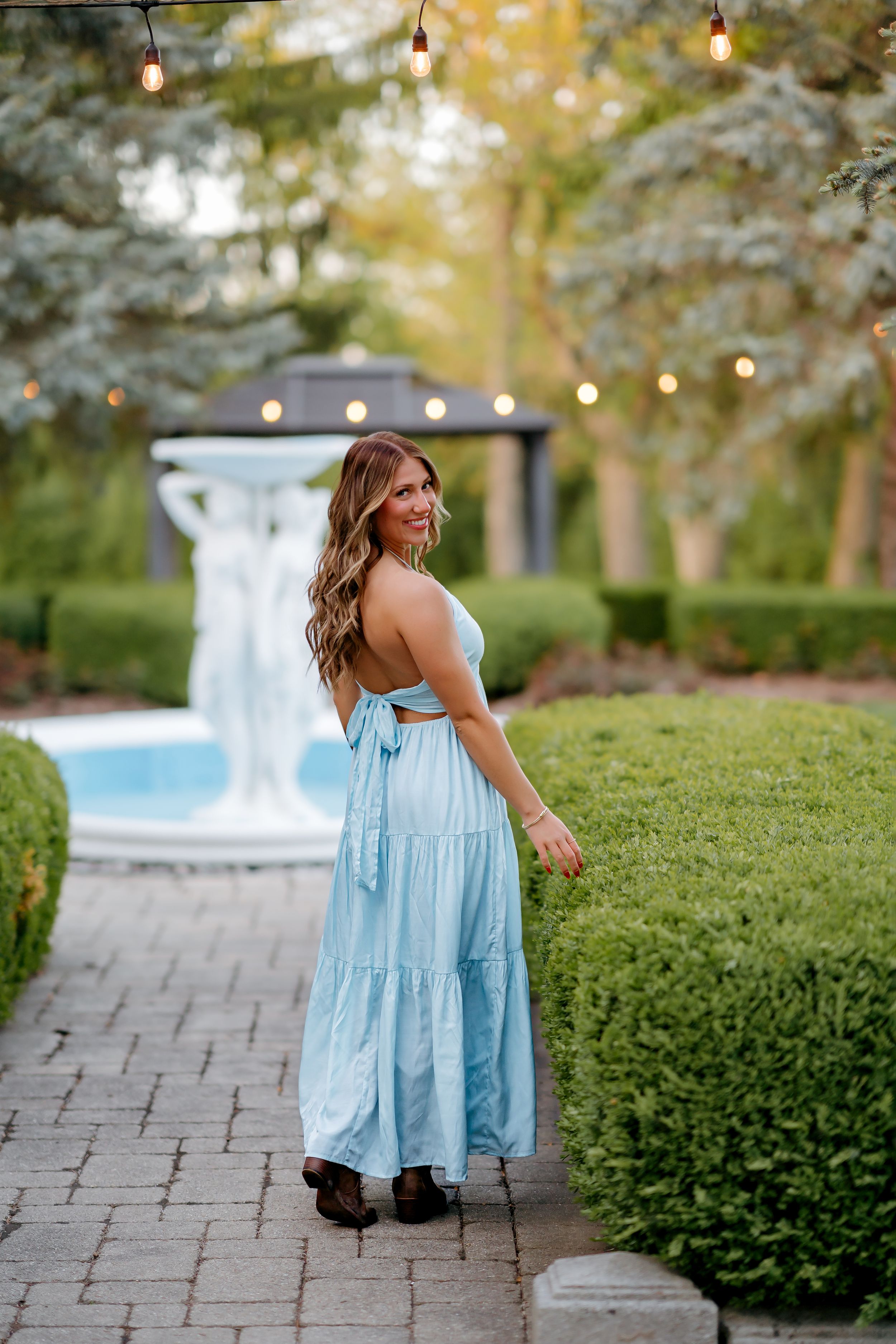 Woman in light blue tiered maxi dress poses in elegant garden with fountain and string lights.