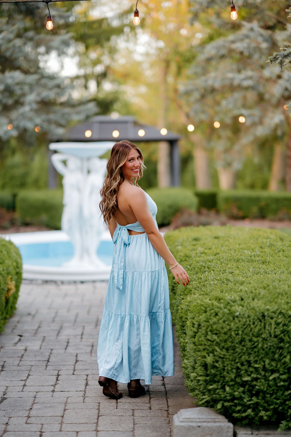 Woman in light blue tiered maxi dress poses in elegant garden with fountain and string lights.