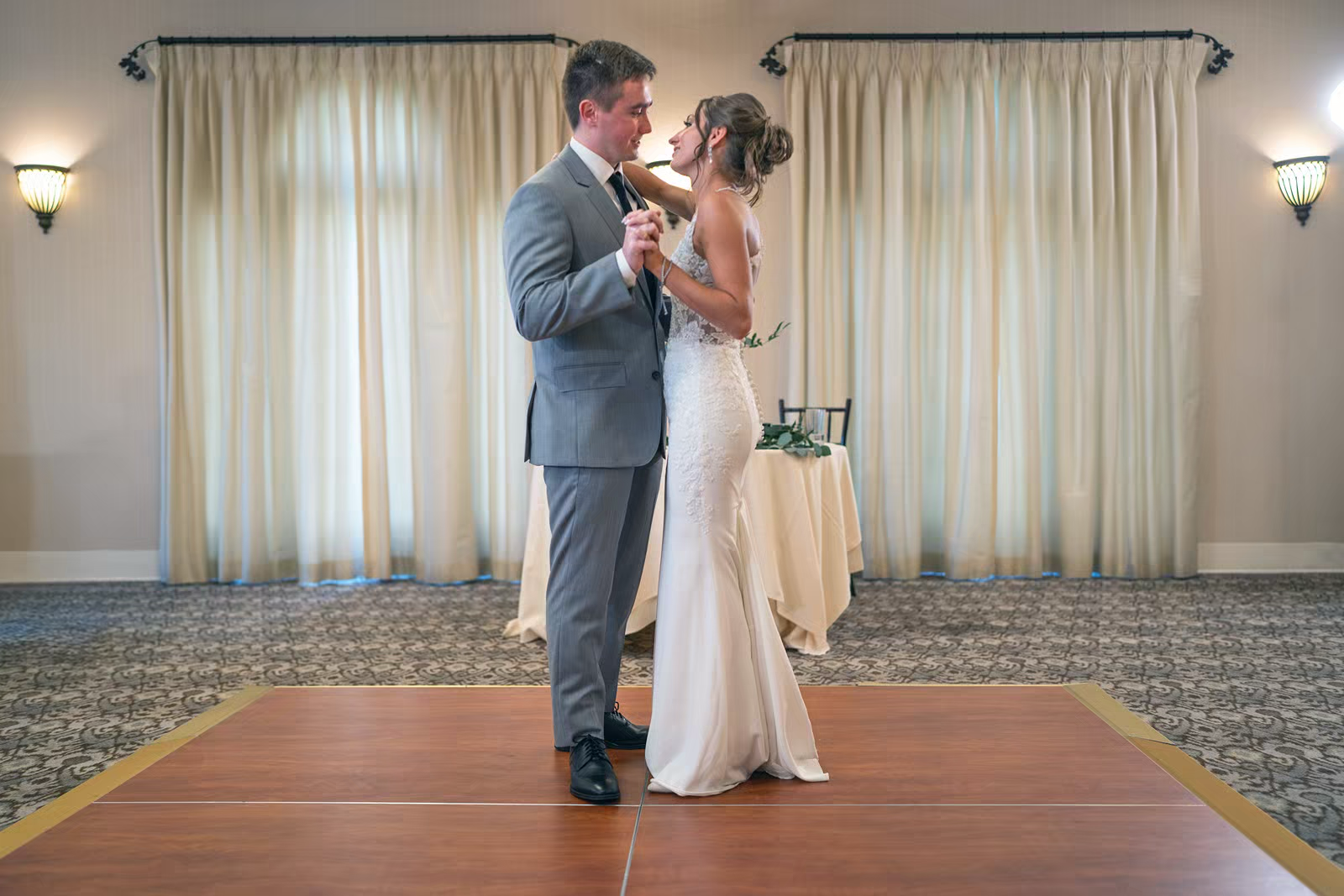 A couple shares their first dance together in a sunlit ballroom with sheer curtains in the background.