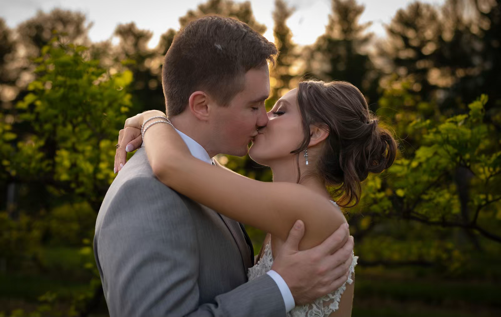 A romantic wedding couple shares a tender kiss outdoors during golden hour with lush greenery in the background.