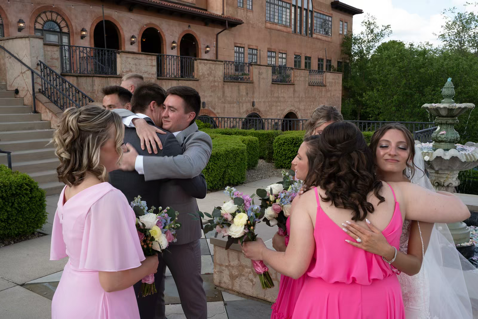 Wedding party in pink bridesmaid dresses and dark suits embrace and celebrate on outdoor patio near brick building.