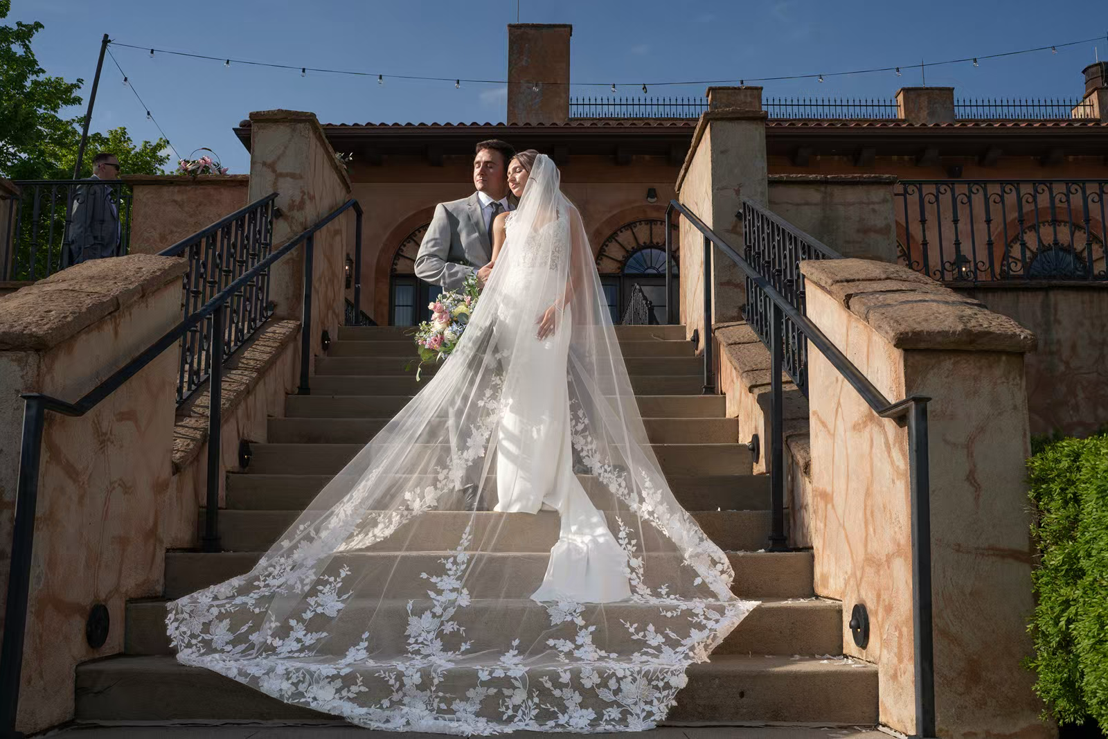 A long lace veil trails down elegant stone steps at an outdoor wedding venue.