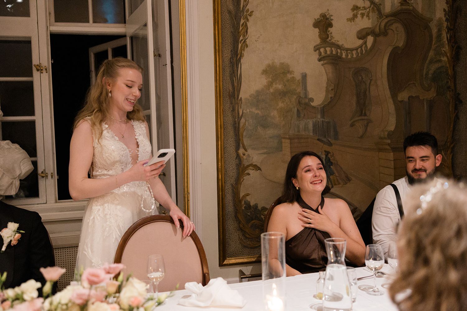 Wedding reception guests seated at an elegant table with decorative wall murals checking their phones during dinner.