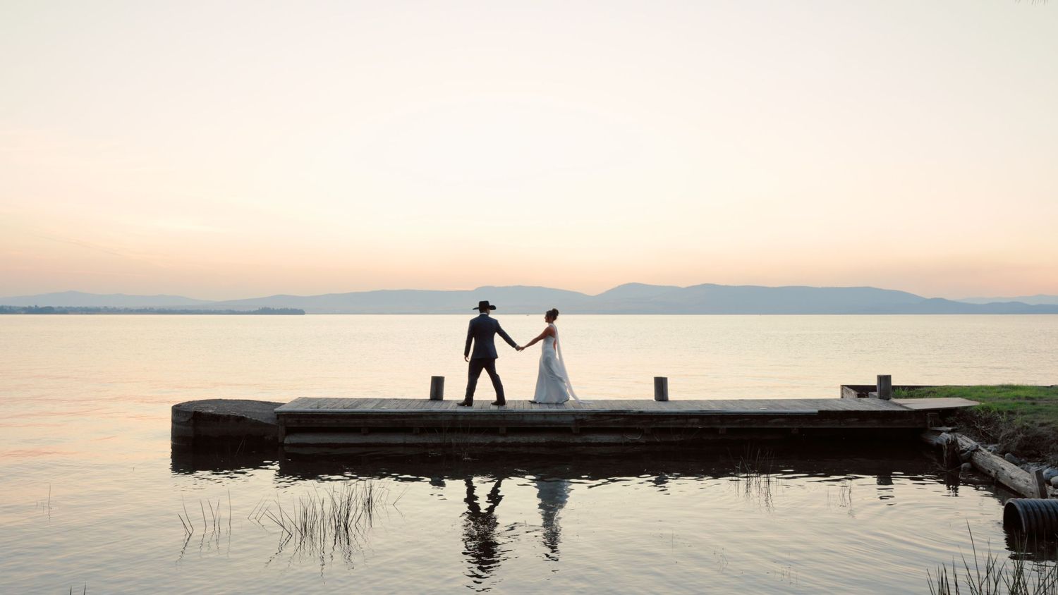 Two silhouettes hold hands while walking through shallow water at sunset along a tranquil lake shore.