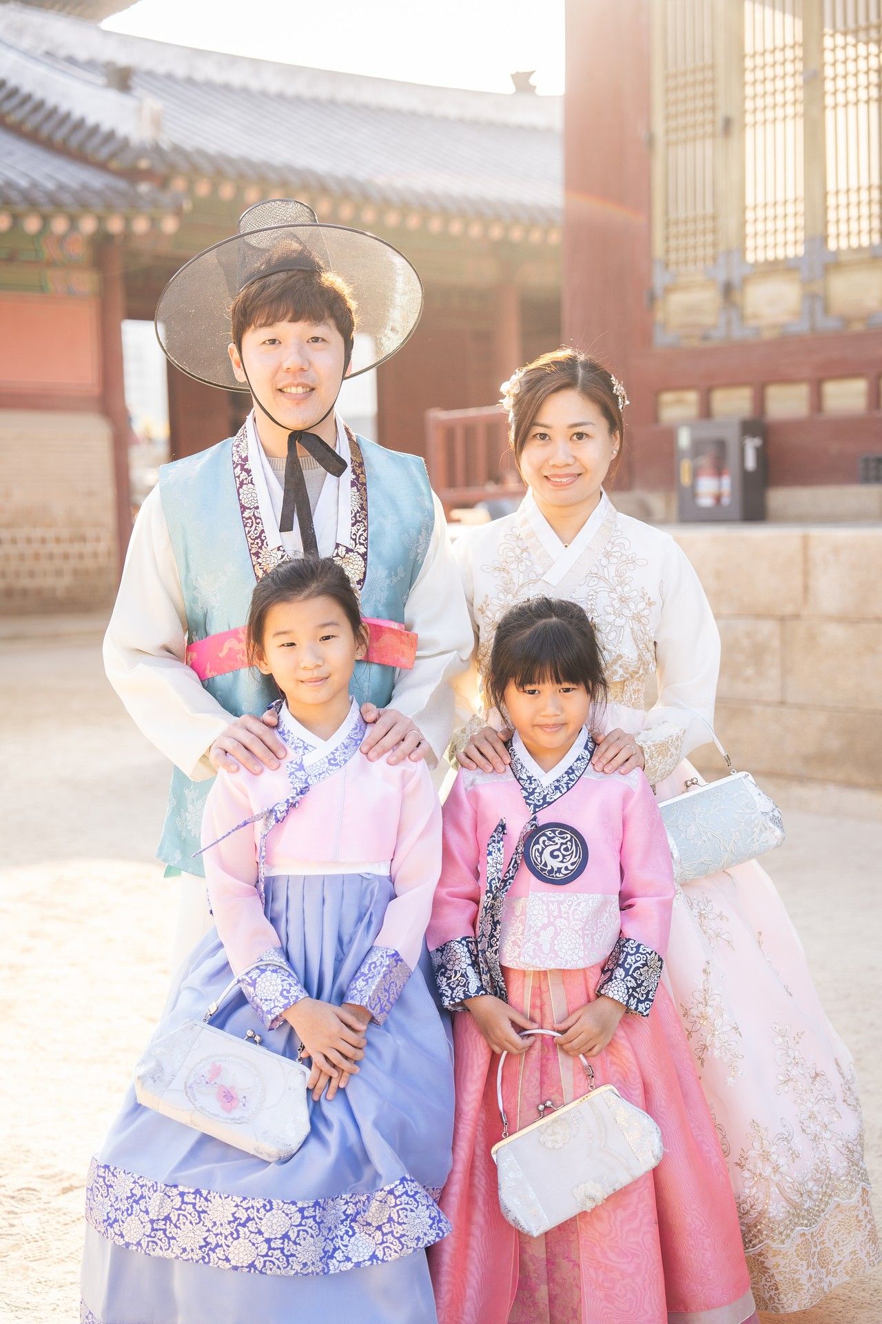 A family wearing traditional Korean hanbok clothing poses together at a historic palace courtyard in bright sunlight.