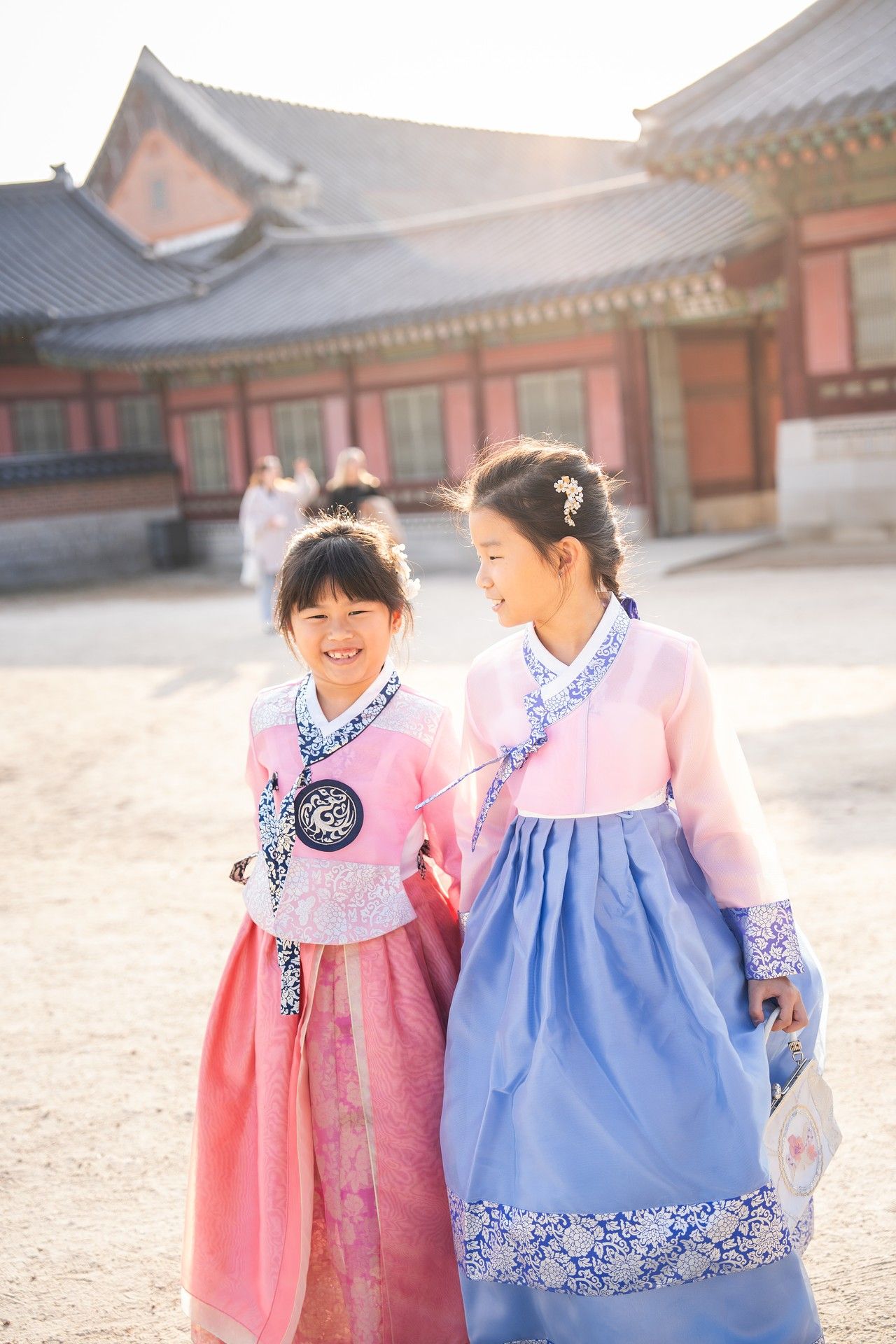 Two people wearing traditional Korean hanbok dresses in pink and blue walk together at a historic palace courtyard.