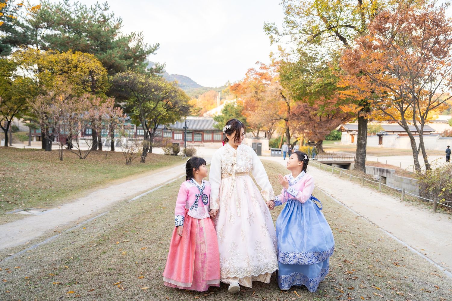 Three people in traditional Korean hanbok dresses walk along a path surrounded by autumn trees with golden foliage.