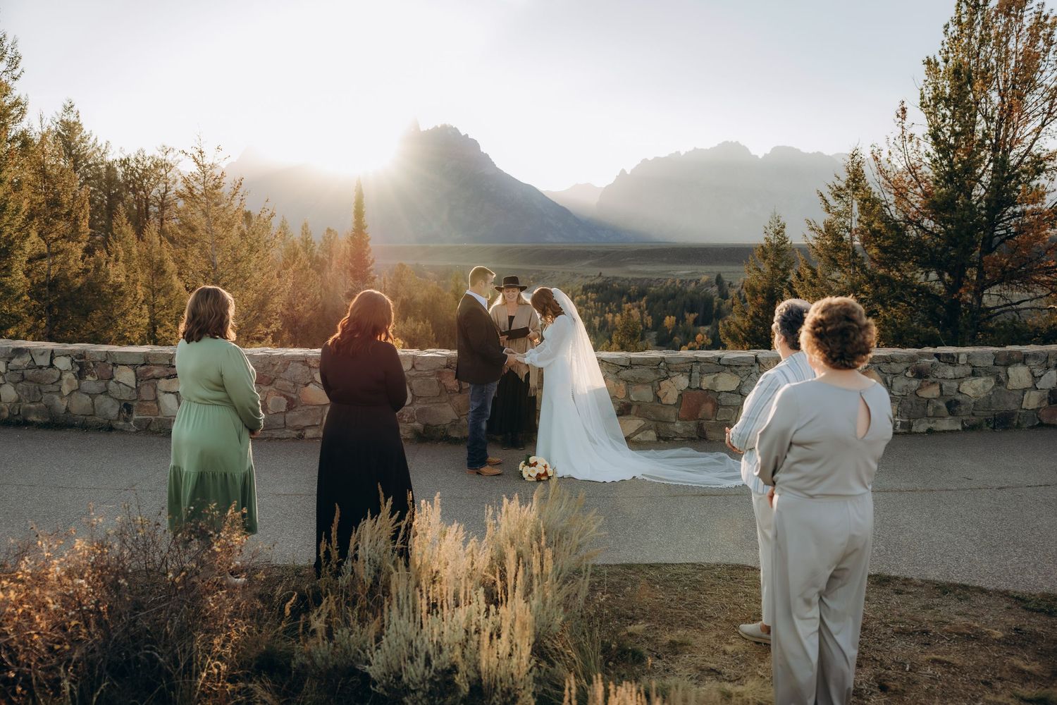Couple in wedding attire stands together against a mountain backdrop at sunset during their outdoor ceremony.