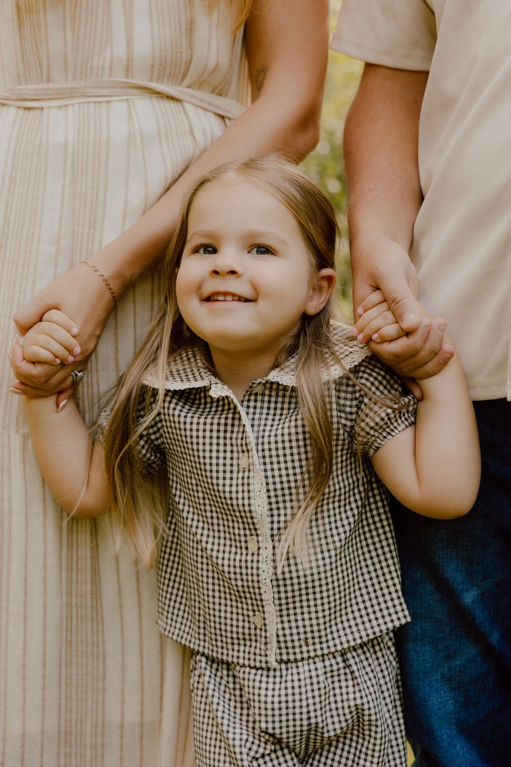 A small child in a checkered dress holds hands with an adult against a light colored background.
