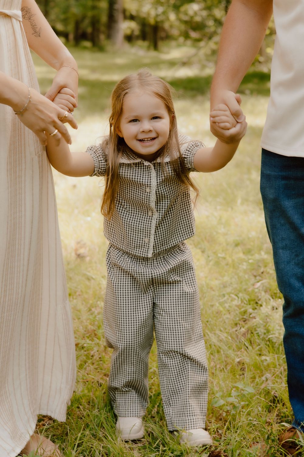 A small child wearing a gingham outfit holds hands with parents while standing in a grassy outdoor setting.