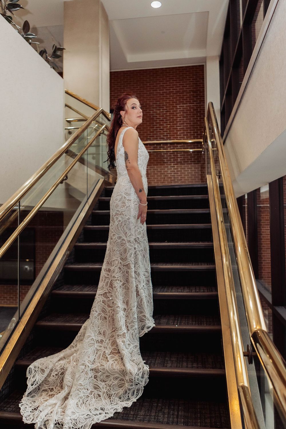 Elegant lace wedding dress with flowing train showcased on staircase with gold railings and brick wall backdrop.