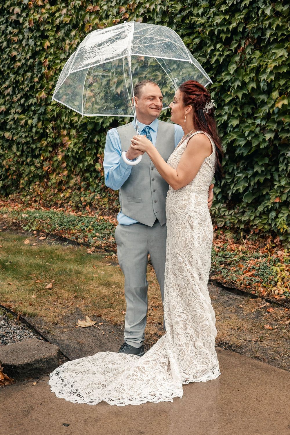 A couple shares a romantic moment under a clear umbrella on a rainy wedding day with ivy-covered walls in the background.