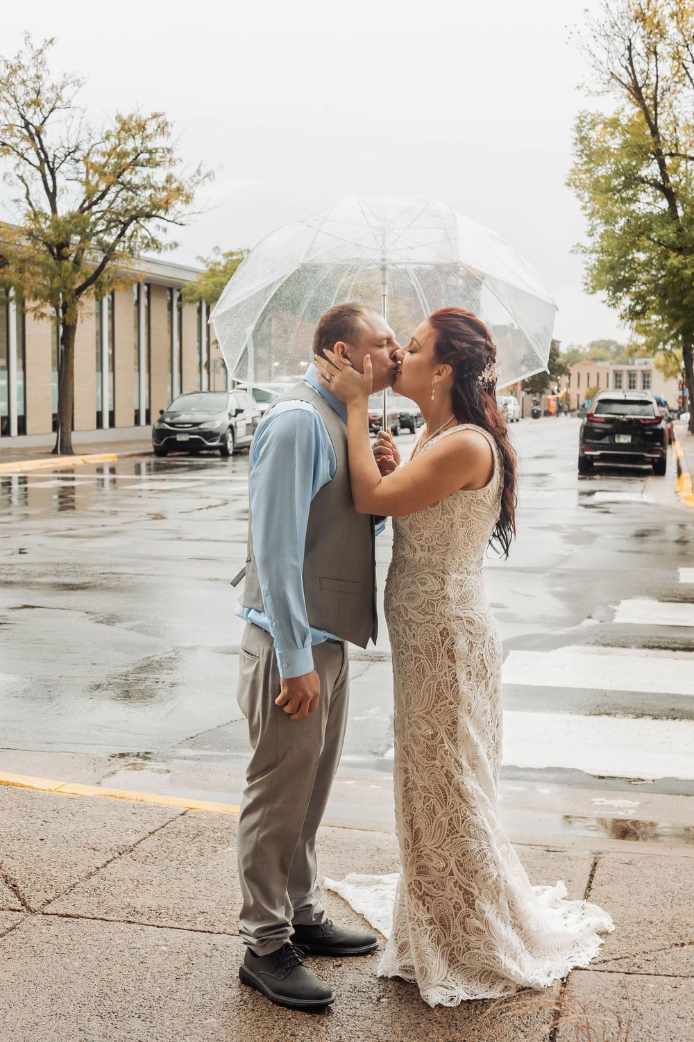 A couple shares a romantic kiss in the rain under clear umbrellas, with the bride wearing a lace gown and groom in gray.
