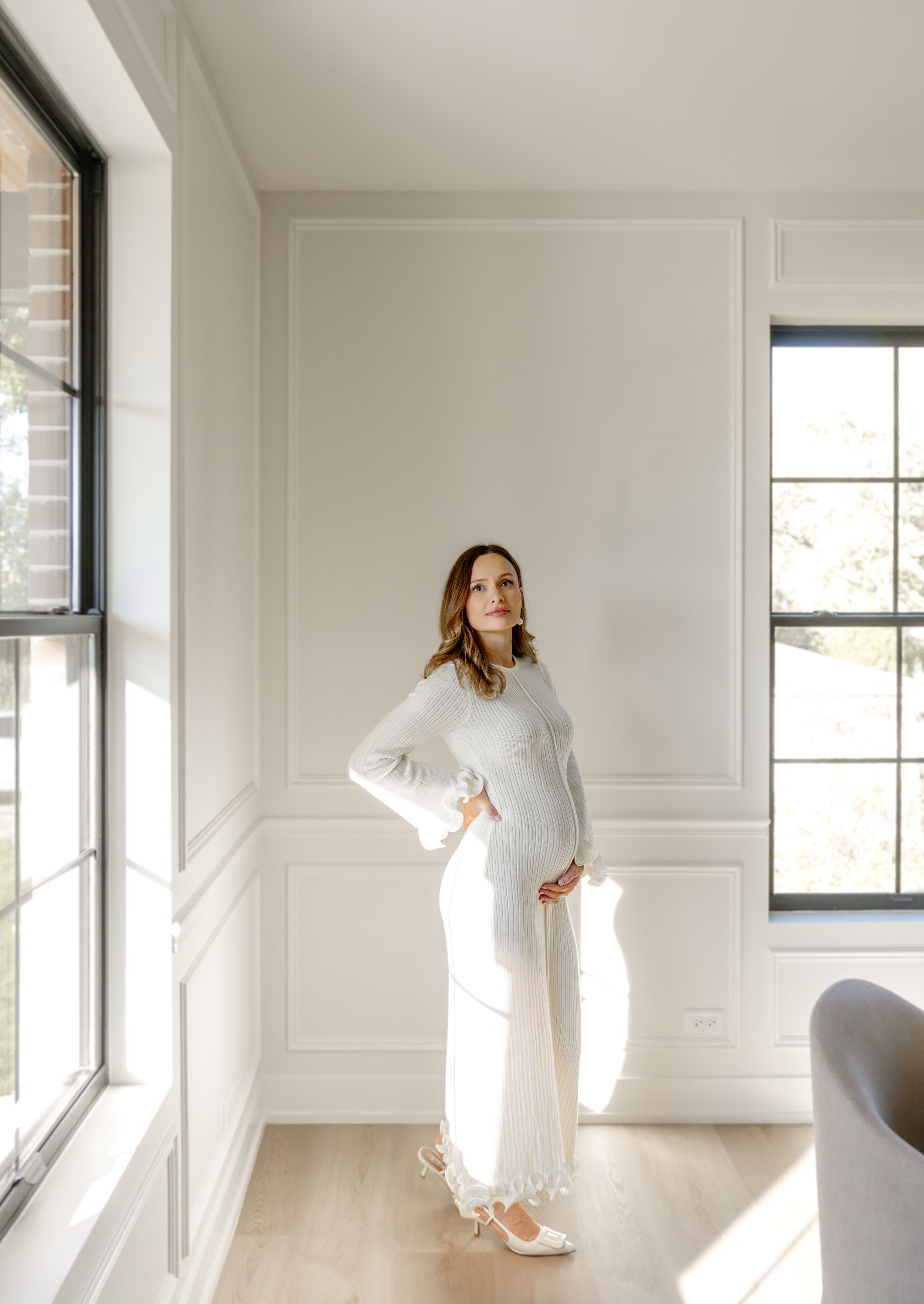 A figure in a flowing white dress moves gracefully through a bright, minimalist room with white walls and windows.