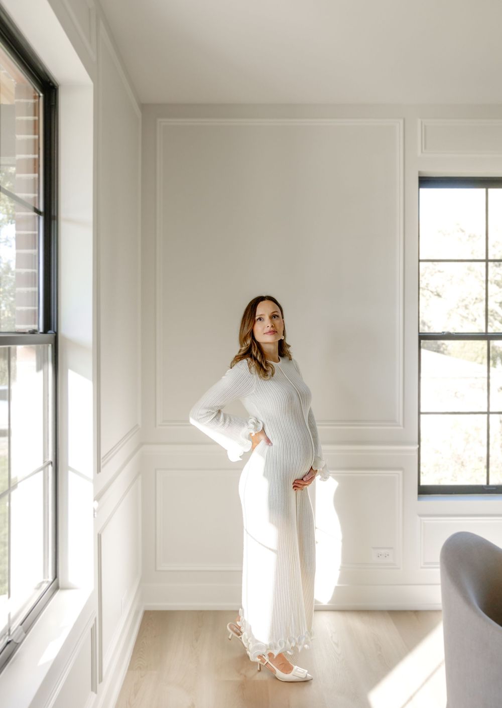 A figure in a flowing white dress moves gracefully through a bright, minimalist room with white walls and windows.