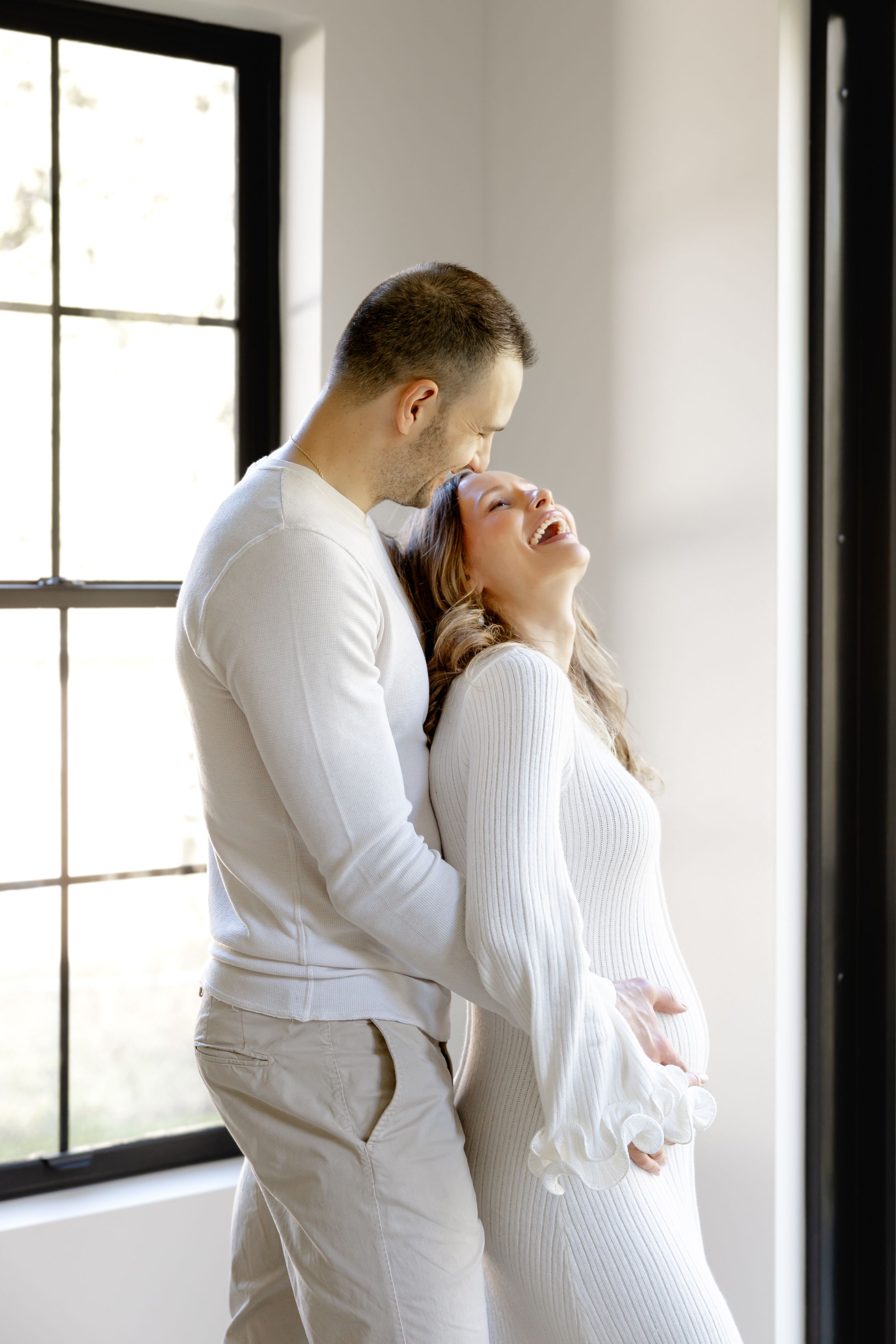A romantic couple dressed in white share a joyful moment together by a bright window in an intimate indoor setting.