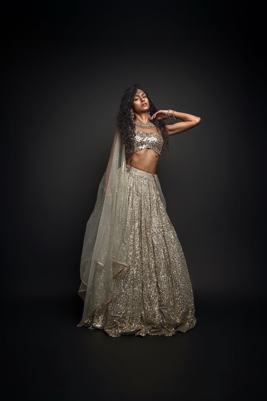 A model poses gracefully in a flowing champagne sequined gown against a dark studio backdrop.