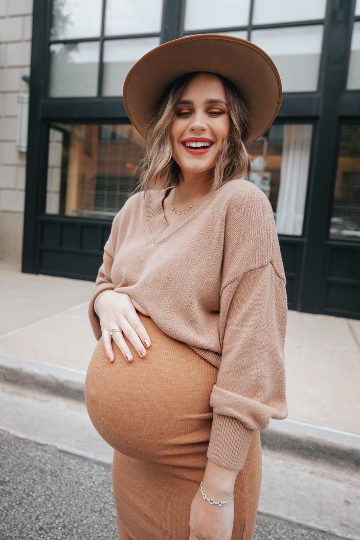 Stylish maternity outfit featuring a camel-colored sweater dress and wide-brim hat in an urban setting.