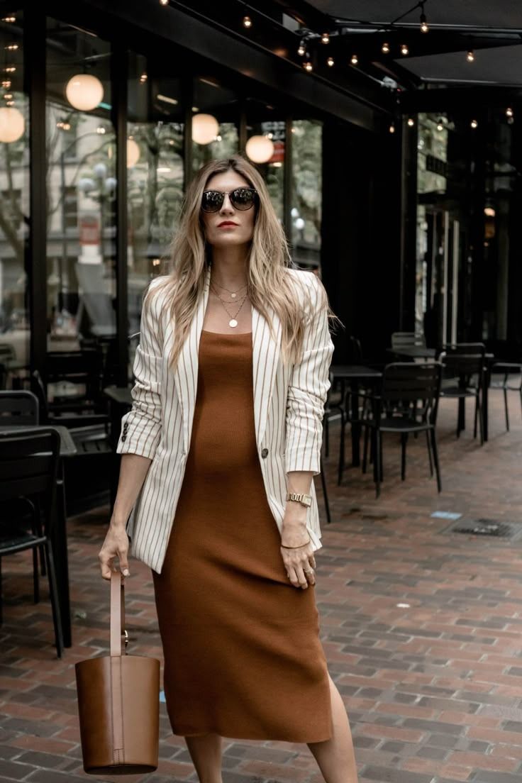 Fashionable outfit featuring brown midi dress with striped blazer and bucket bag on brick patio with string lights overhead.