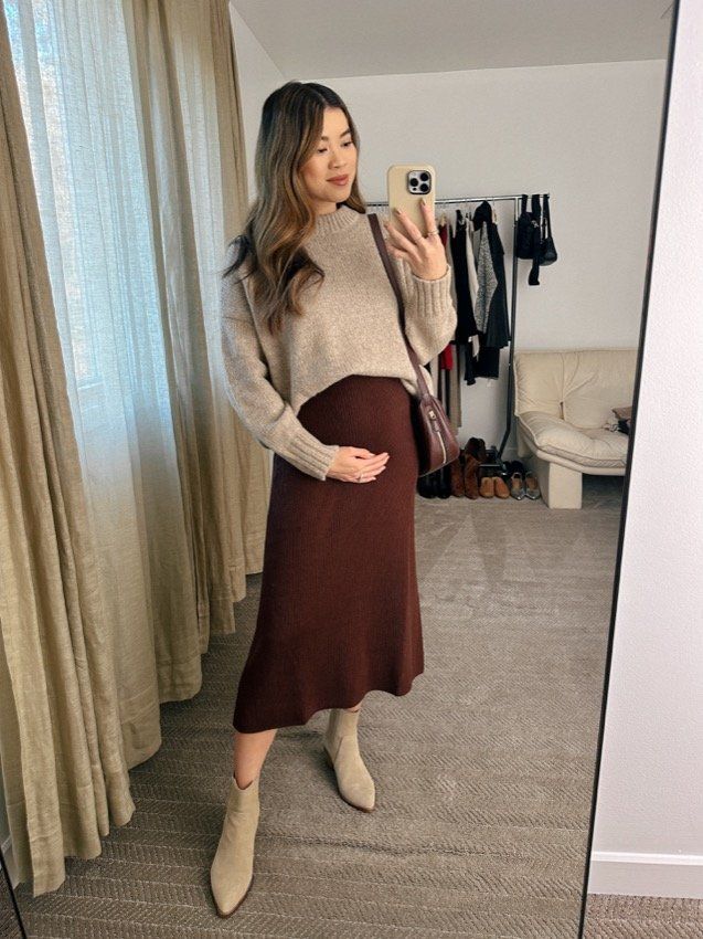 Mirror selfie of an outfit featuring a beige sweater with brown skirt and light colored boots in a bedroom.