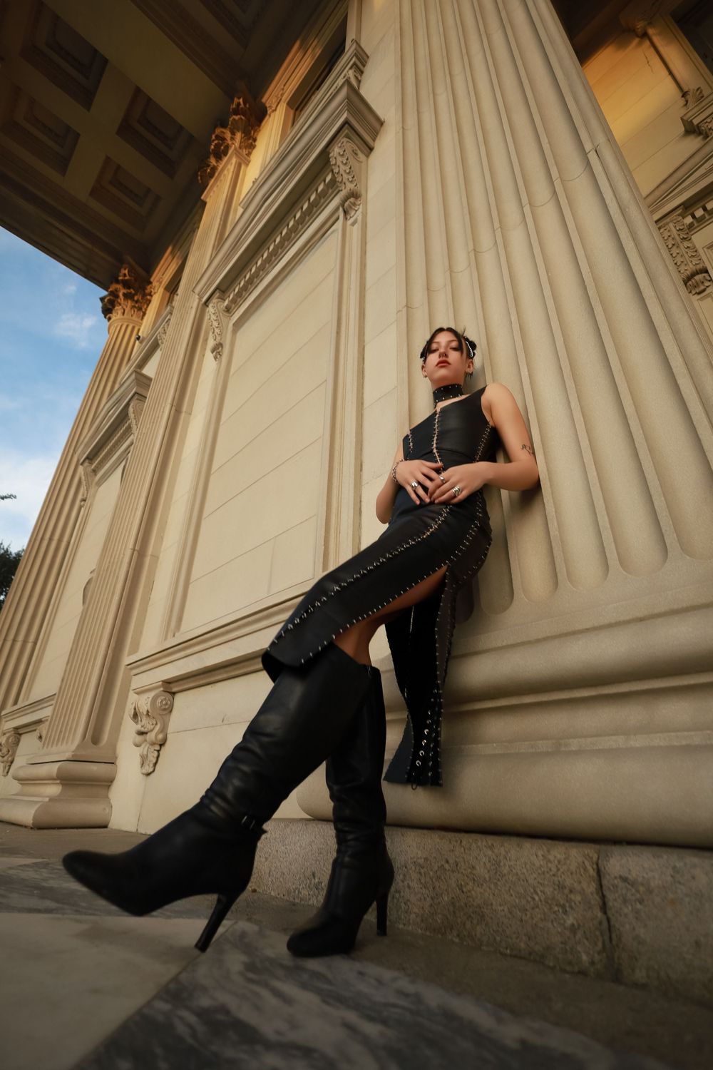 Gothic fashion model poses dramatically against classical stone columns in a striking black outfit and boots.