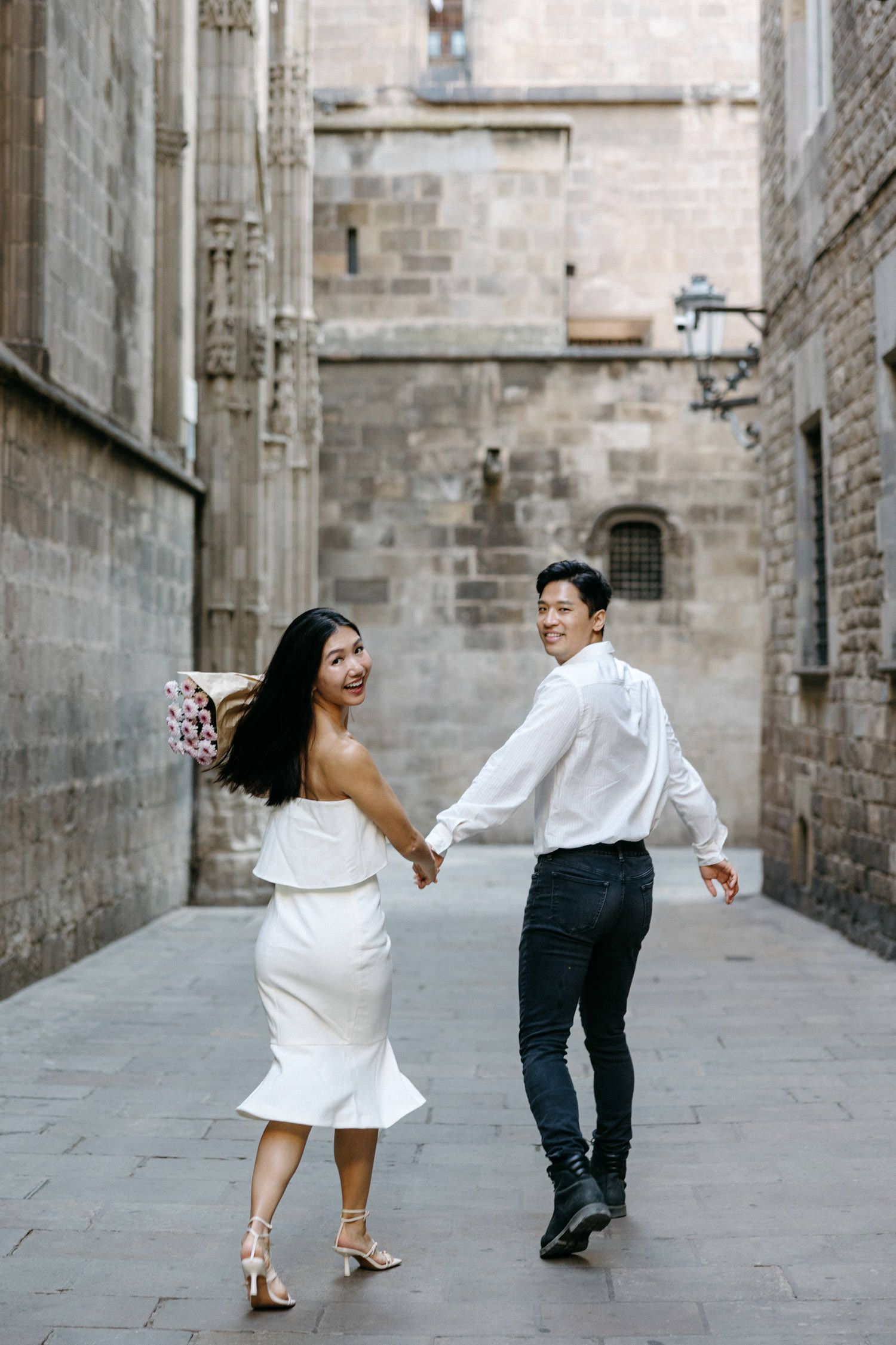 Couple in white dress and dark outfit dancing playfully in a historic stone alleyway.