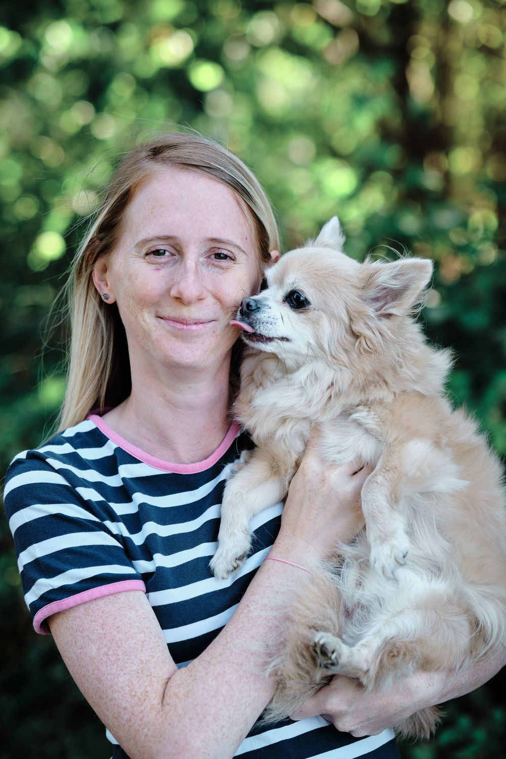 A cream-colored Chihuahua mix dog is held by someone wearing a black and white striped shirt against a green outdoor background.