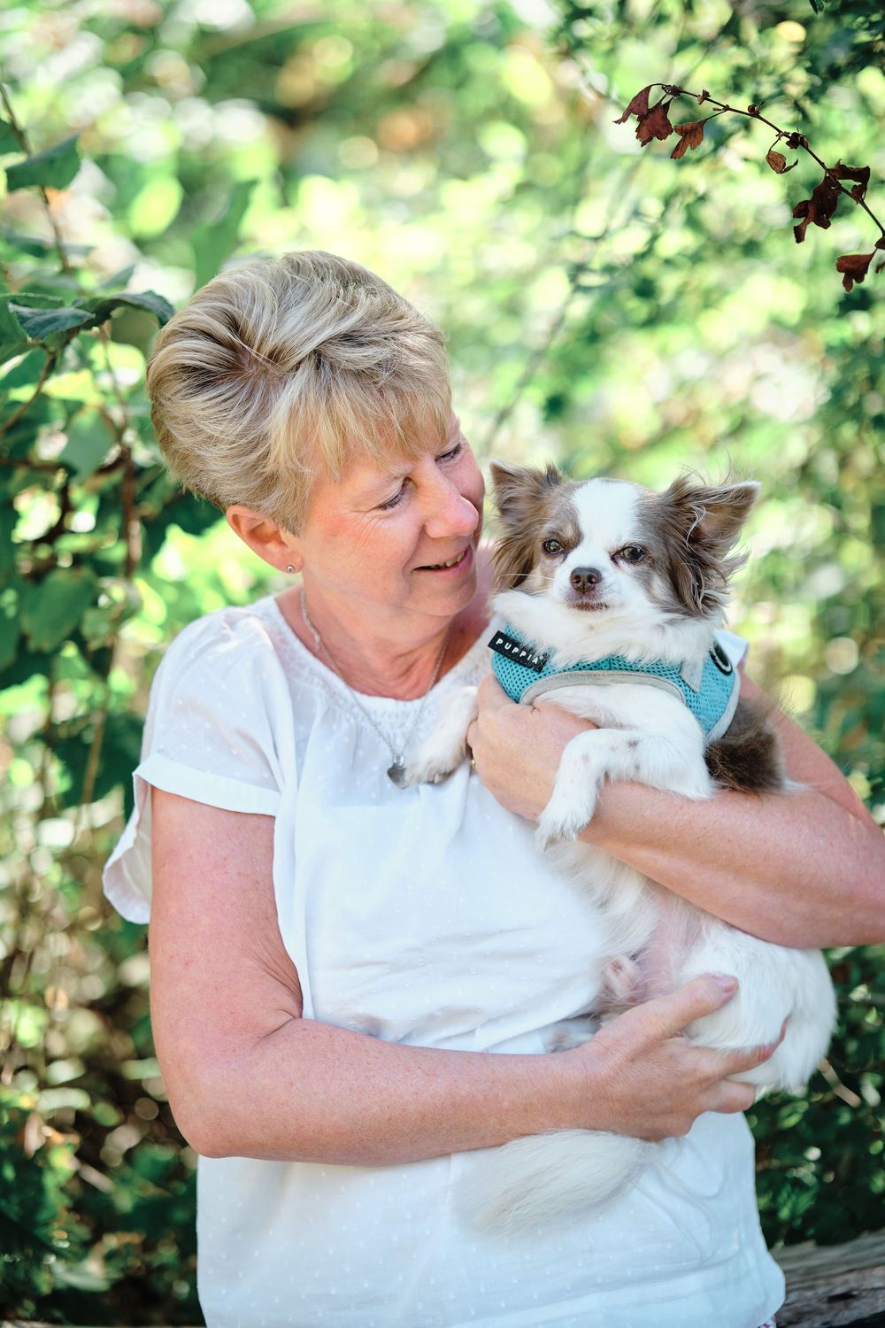 A person in a white shirt holds and cuddles with a small white fluffy dog outdoors against a green leafy background.
