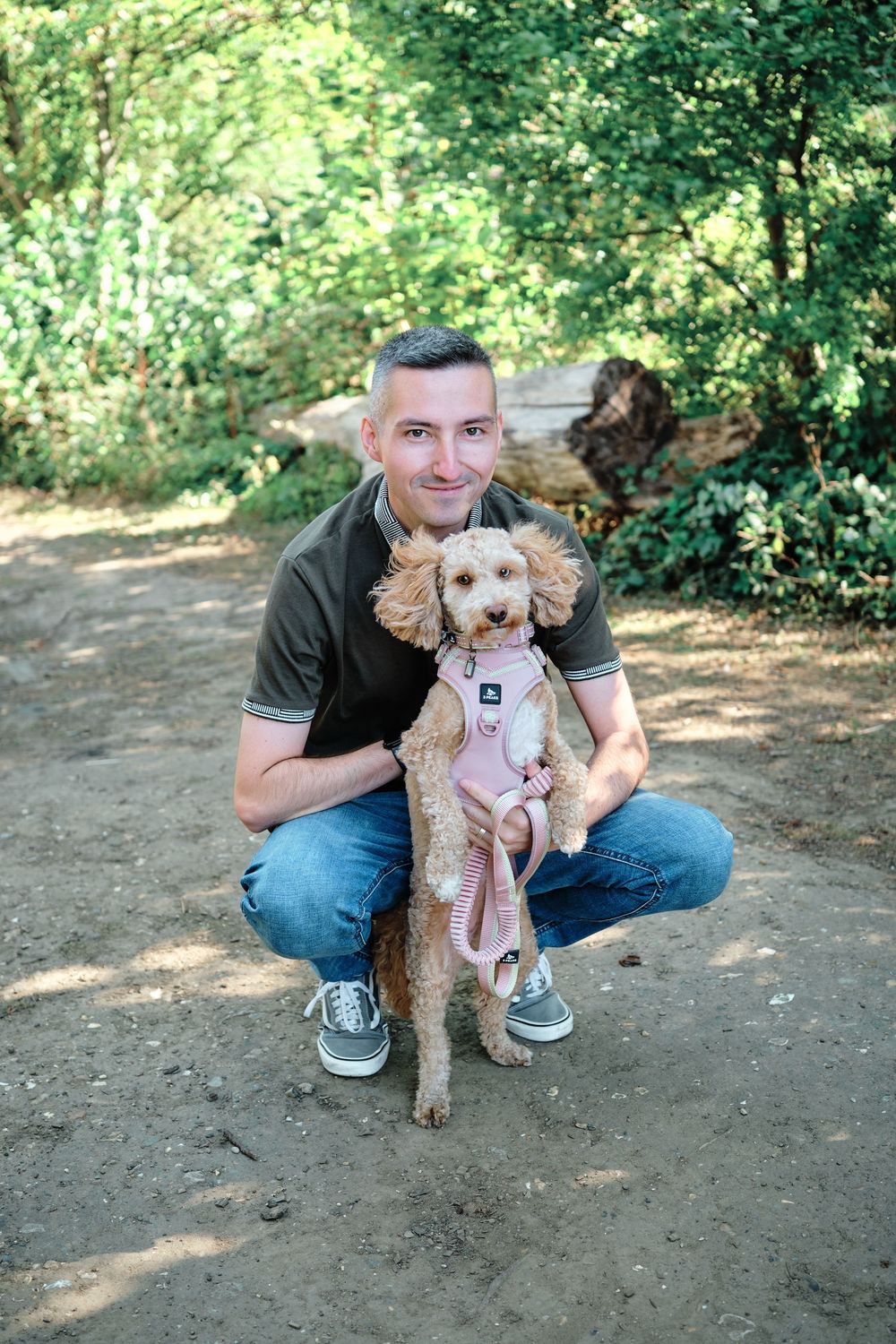 A person crouches on a dirt path in a forest while holding a small goldendoodle dog wearing a harness.