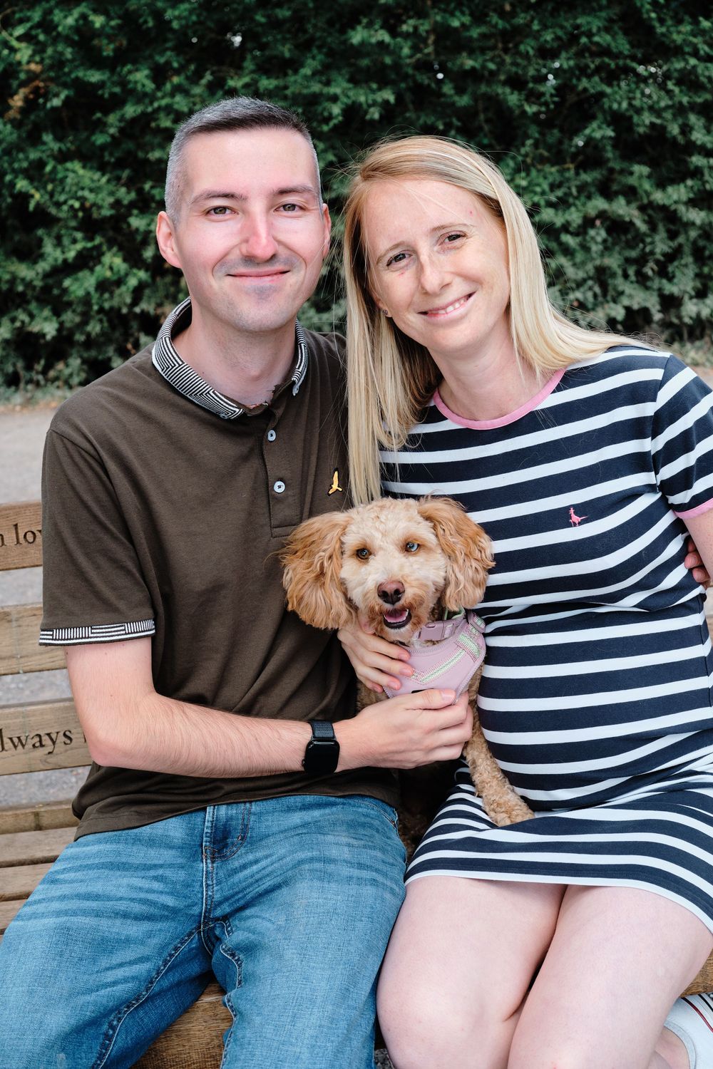 A couple sits on a wooden bench with their small golden-colored dog, wearing casual outfits in an outdoor setting.