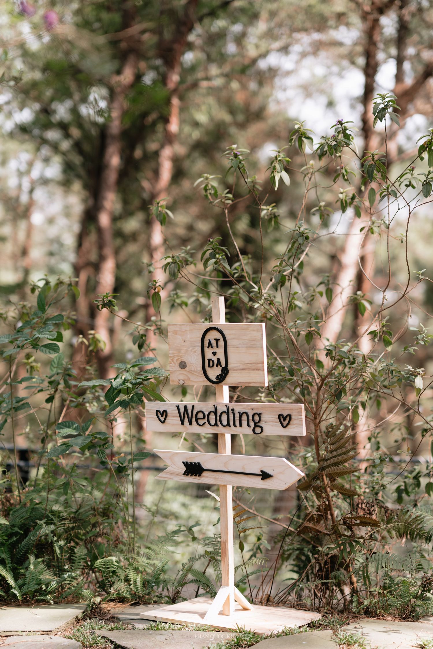 Rustic wooden wedding directional sign with hearts in a sunlit forest setting.