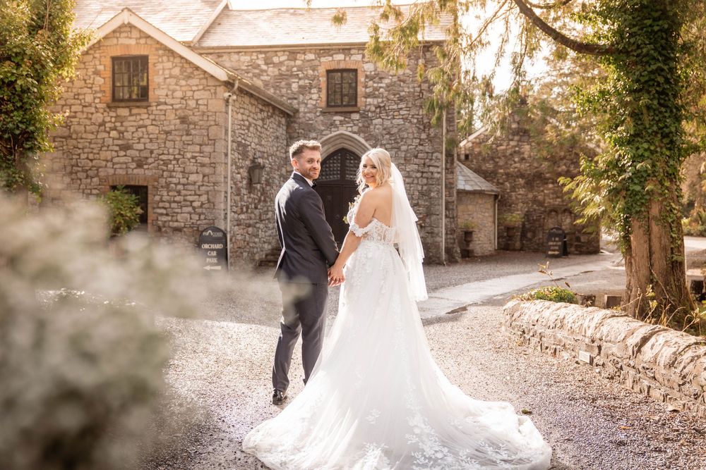 A romantic wedding portrait in front of a rustic stone building with soft natural light filtering through the trees.