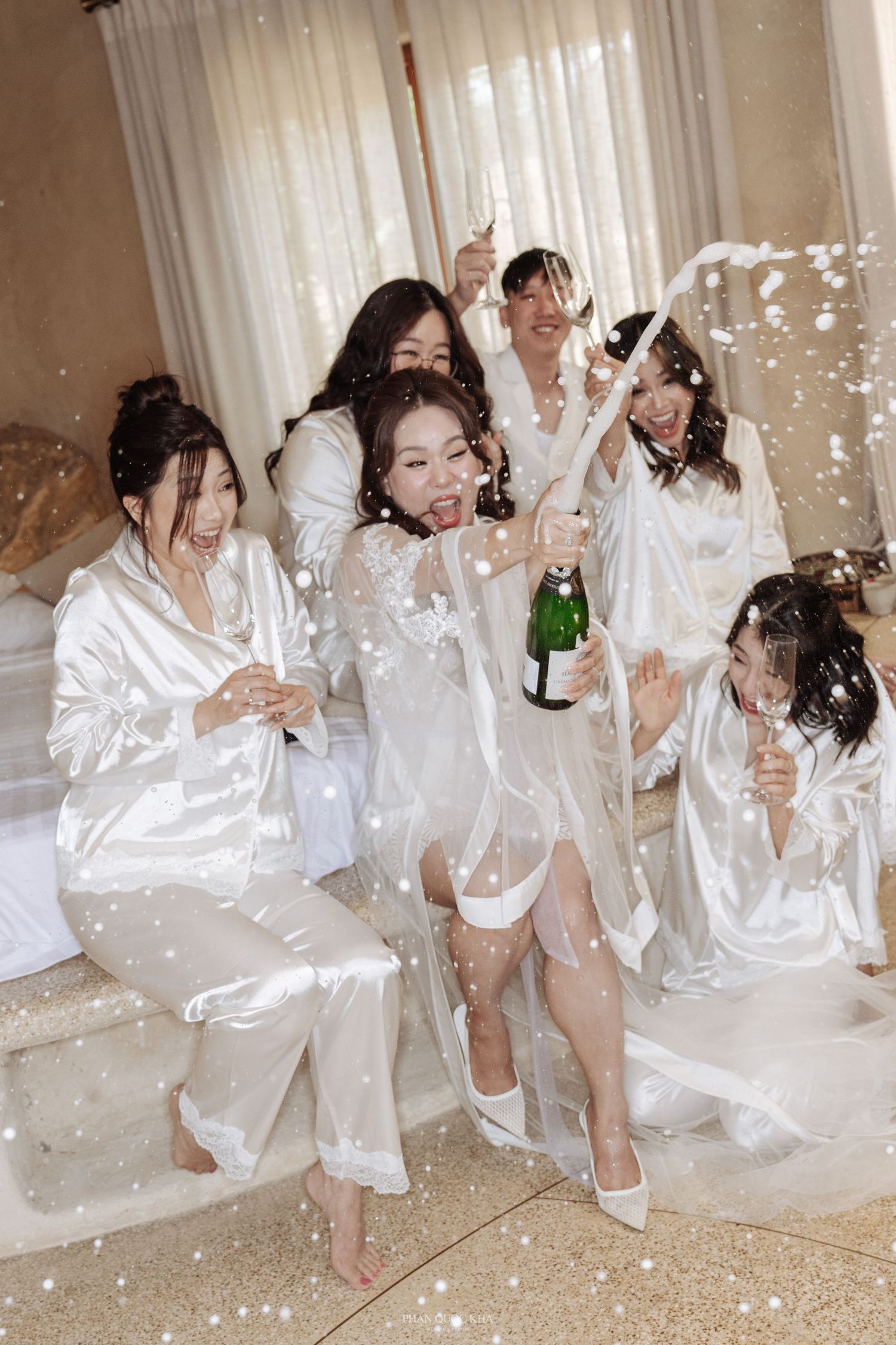 Bridesmaids in white satin robes celebrate with champagne and confetti during bridal getting ready party.