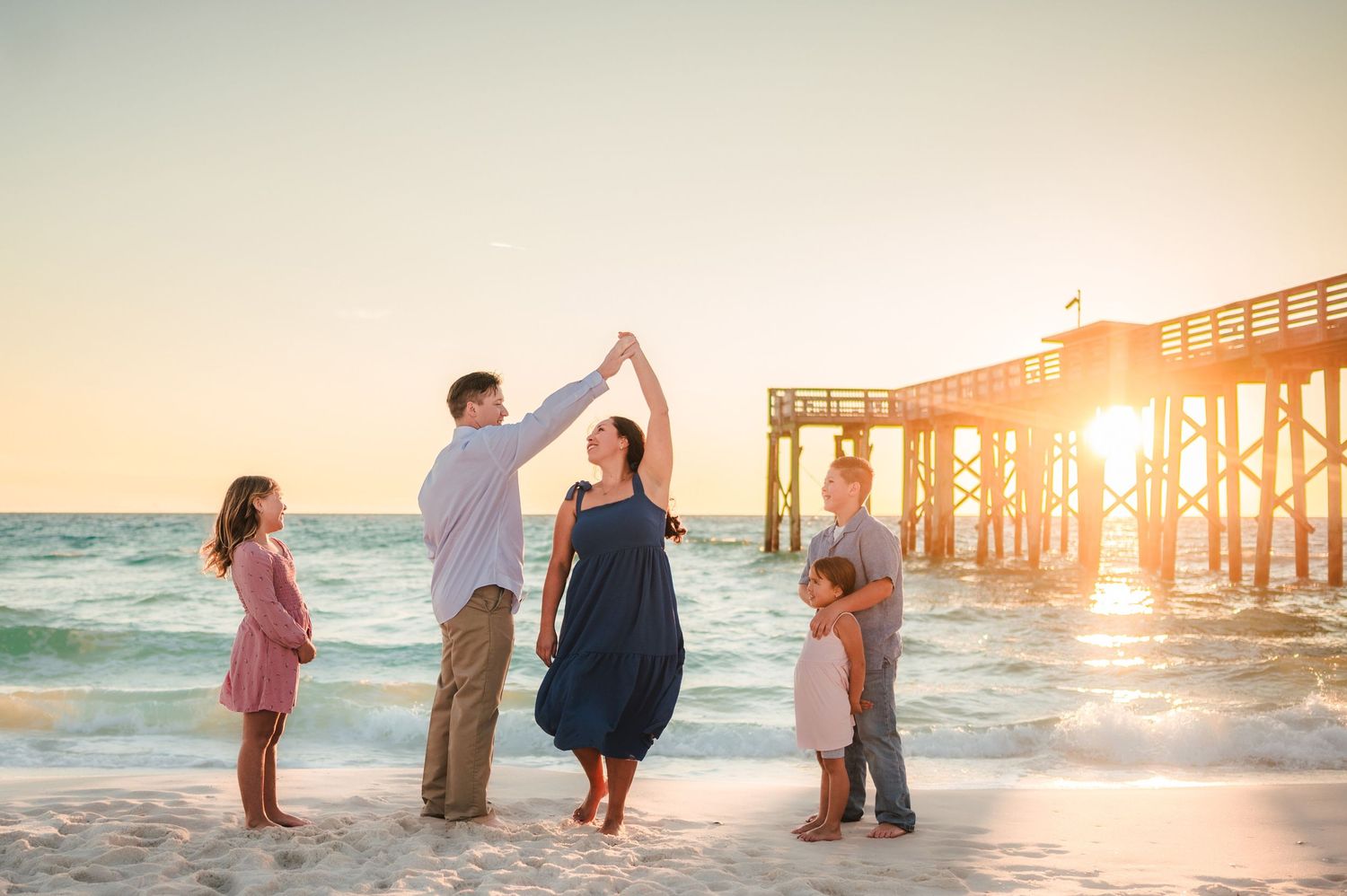 A group stands together on a beach at sunset with a wooden pier in the background.