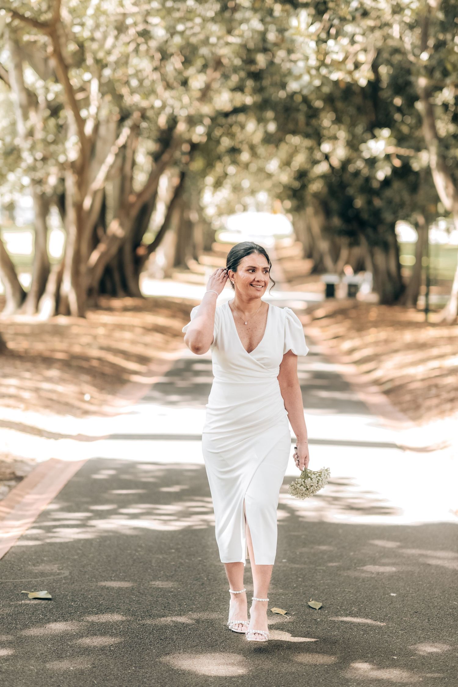 A bride in a white midi dress walks down a tree-lined path on a sunny day holding a small bouquet.