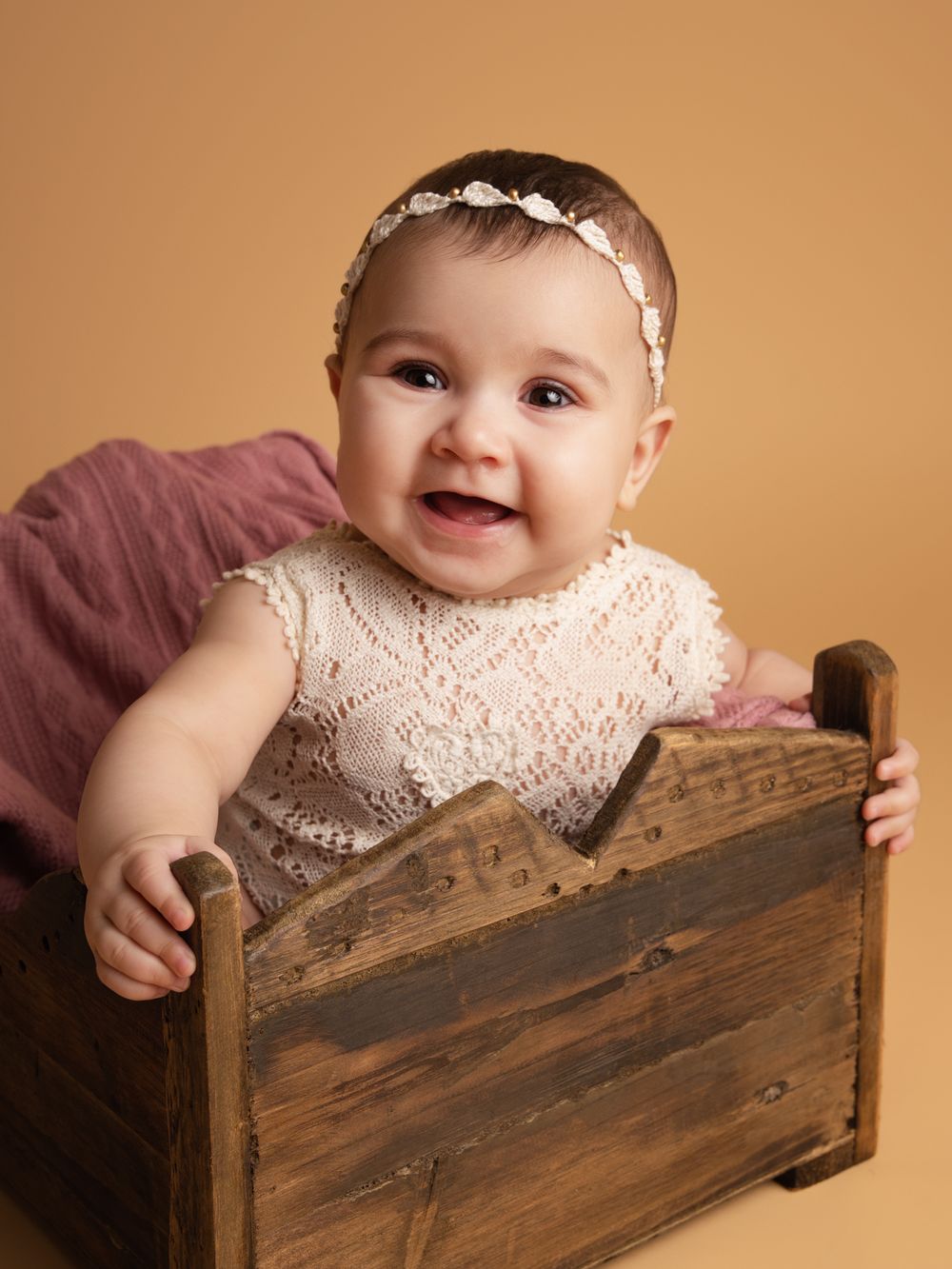 Smiling baby girl in cream lace romper and floral headband sitting in rustic wooden crate studio portrait.