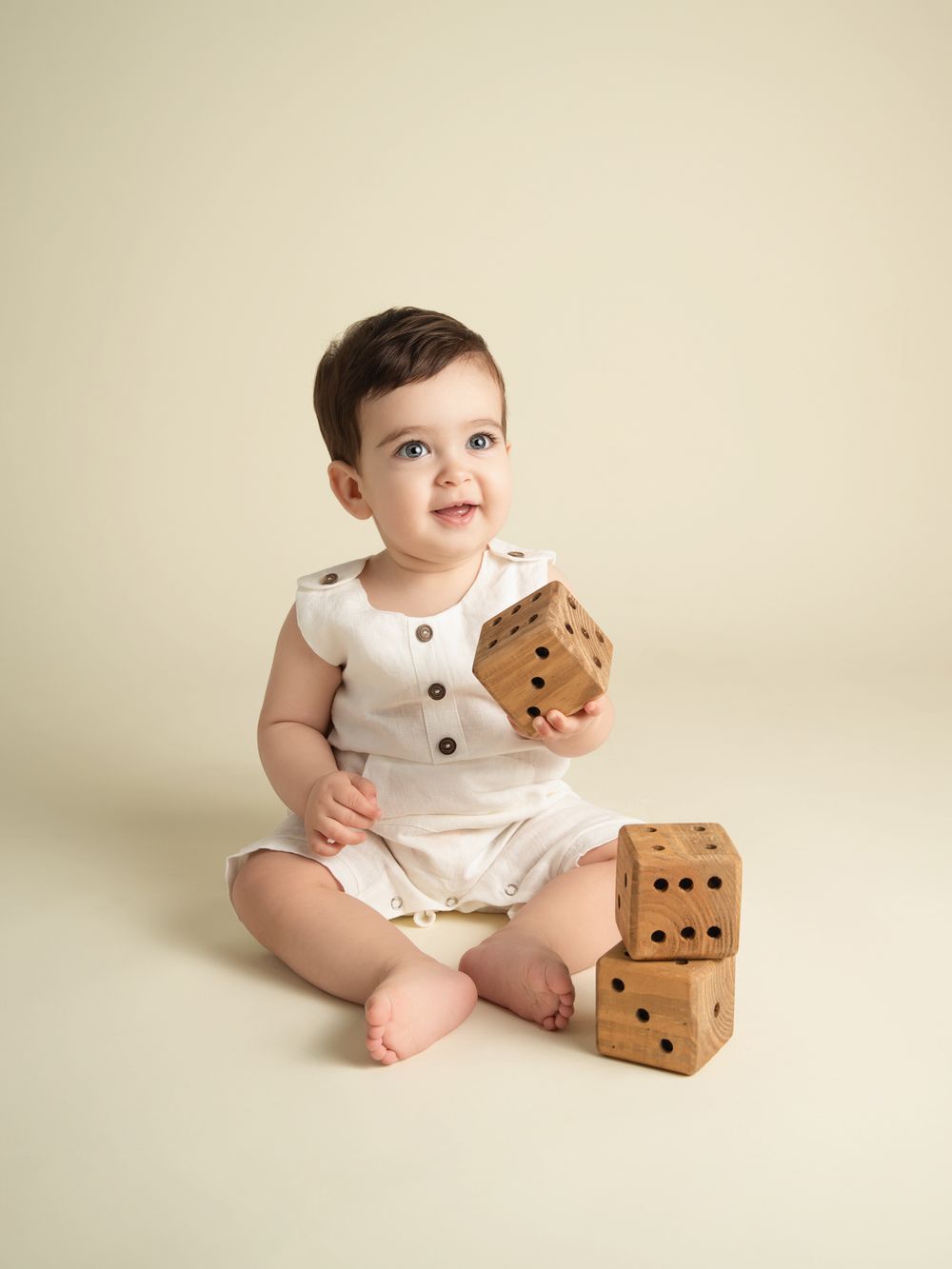Adorable baby in white romper holding wooden dice blocks during milestone photography session.