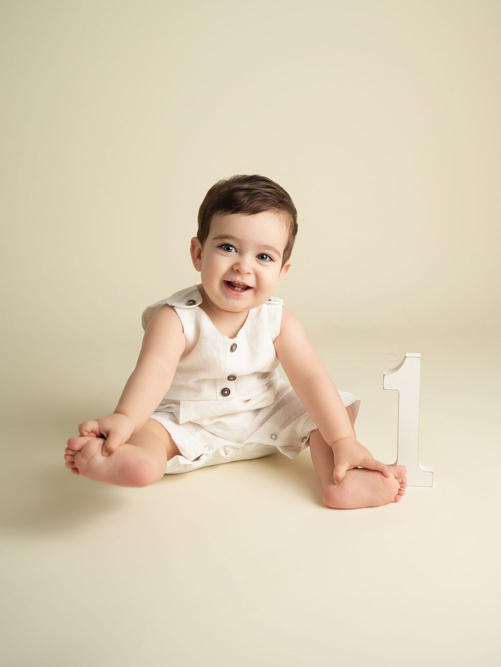 Smiling baby in white romper sitting next to number one sign for first birthday photo session.