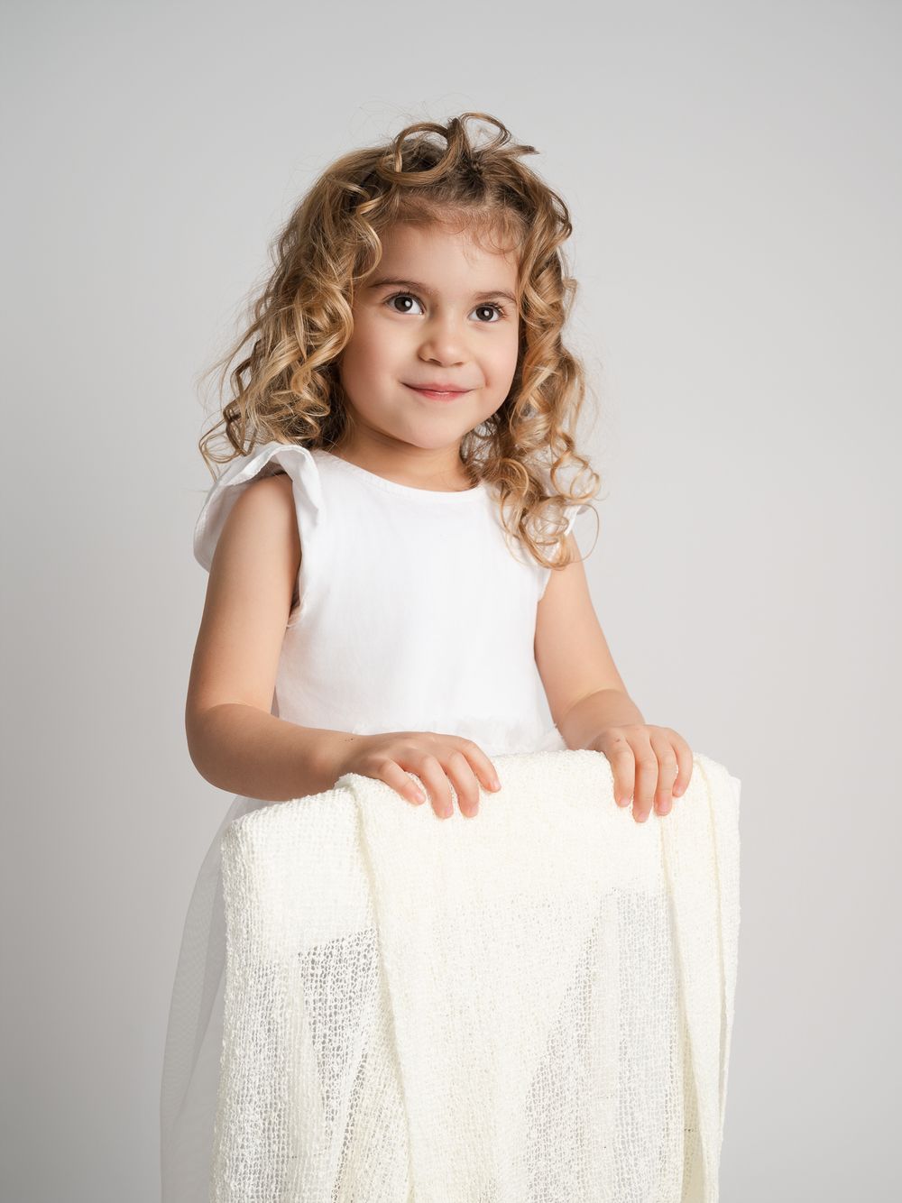 Curly blonde girl in white dress holding tulle in studio.