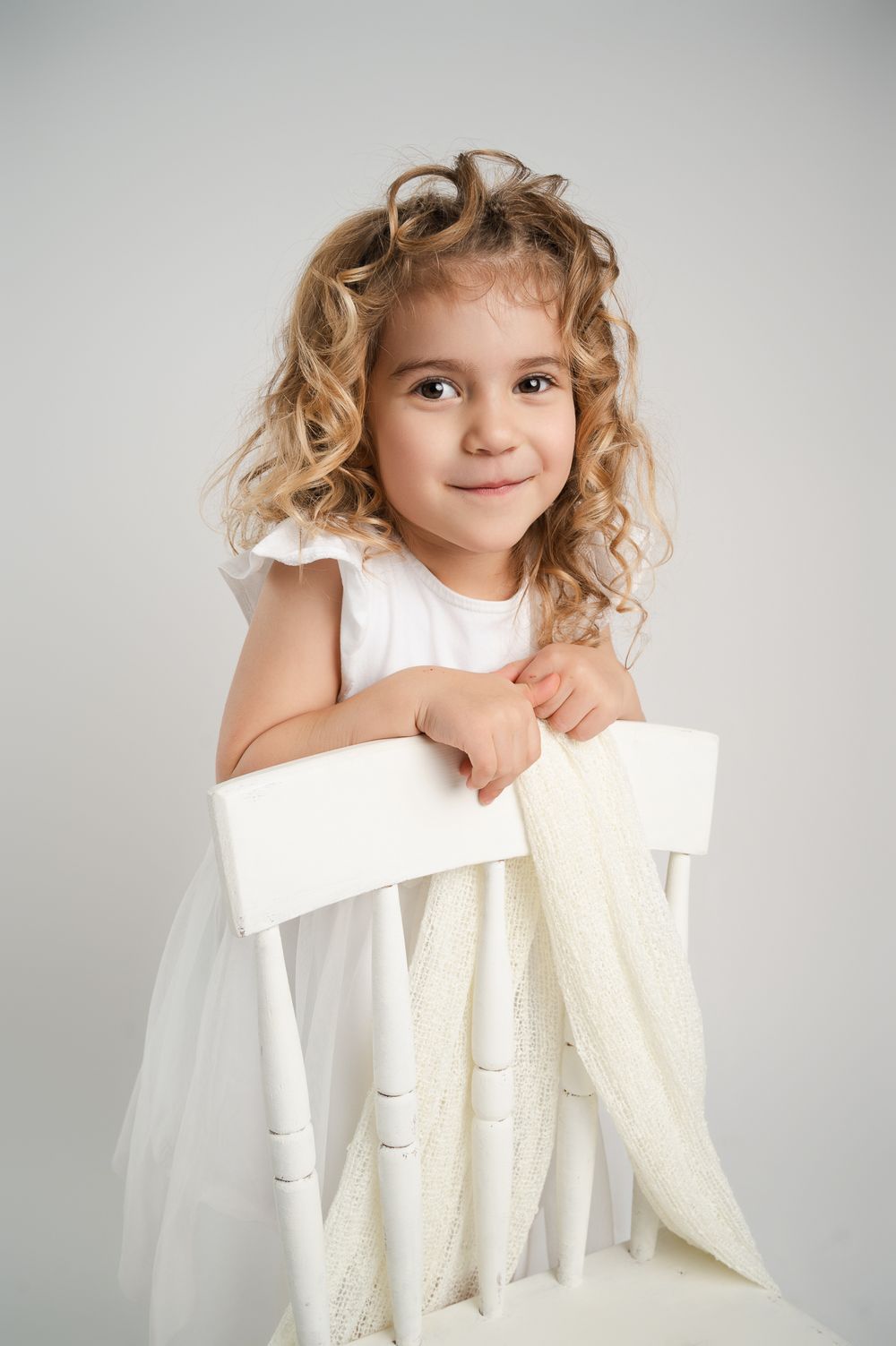 Curly blonde girl in white dress leaning on white chair.