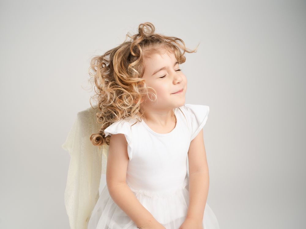 Young girl in white angel dress with wings poses with eyes closed and curly blonde hair in studio portrait.