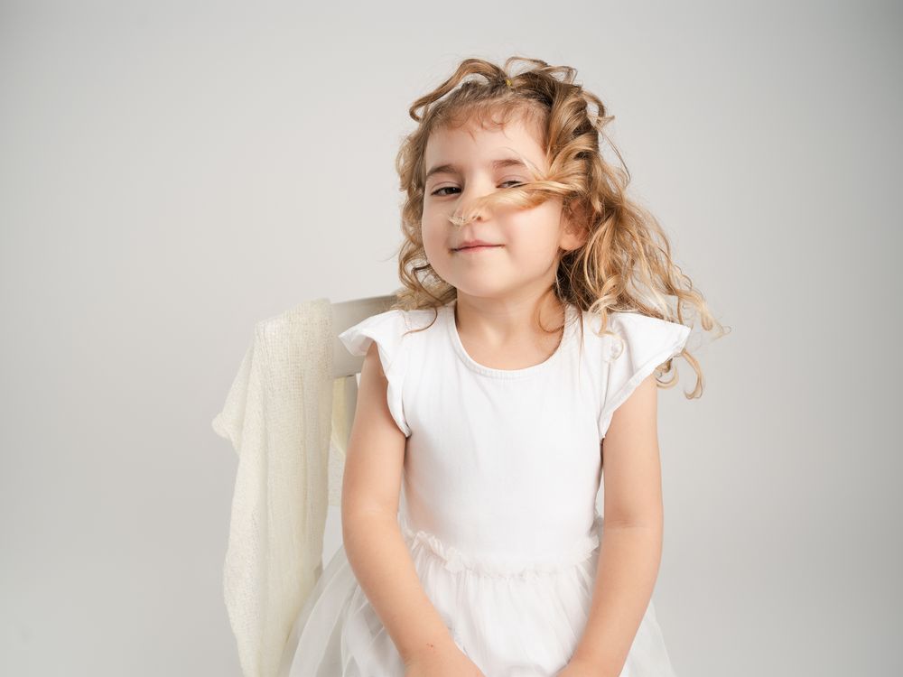 Adorable little girl wearing white angel costume with wings smiles playfully in professional studio photo.