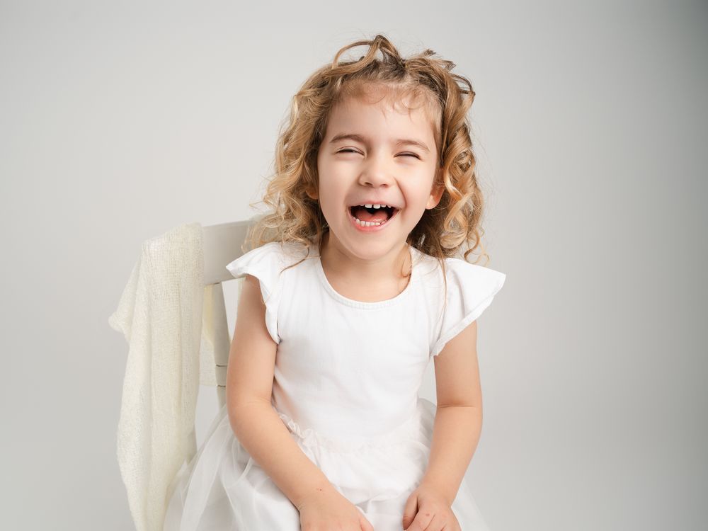Happy little girl in white angel dress laughs joyfully with wings visible in bright studio portrait.