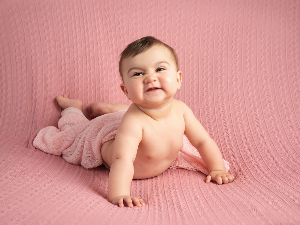 Cheerful baby girl lying on pink knit blanket smiling at camera in soft professional studio portrait.