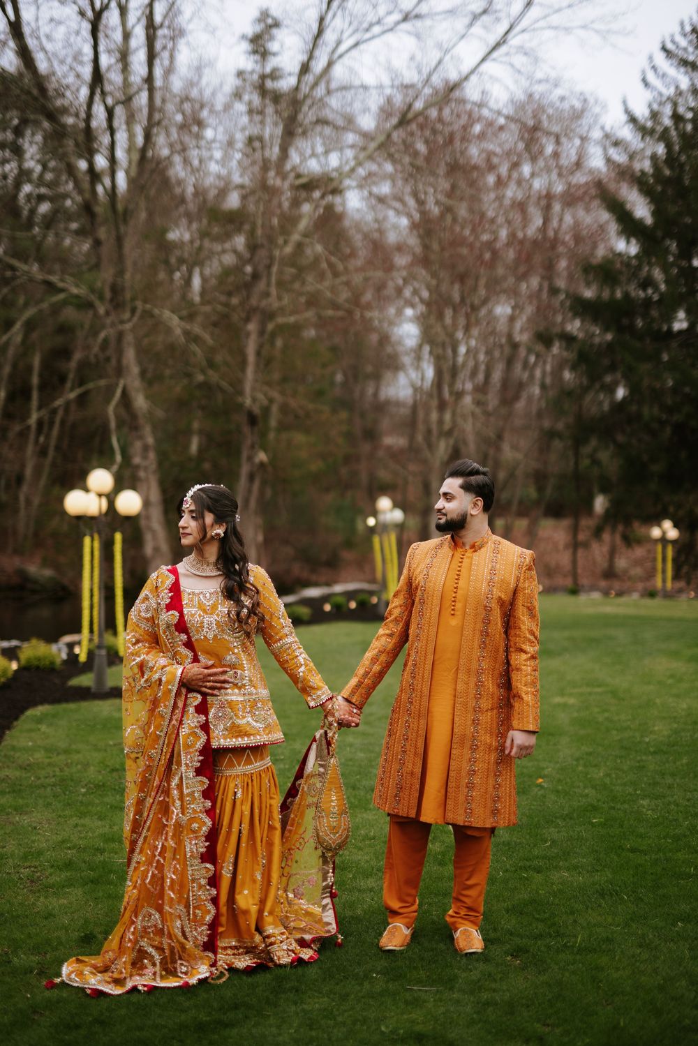 A couple in traditional South Asian wedding attire holds hands while standing on a green lawn with lit lampposts in background.